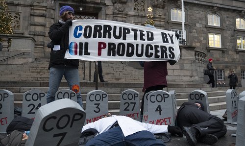 two protestors stand holding a banner that reads corrupted by oil producers while scientists lay on the floor amid fake graves marked with the names and years of all the COP summits