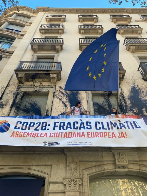 protestors and scientists stand on the black paint splattered balcony of the european union offices with a banner that reads cop28 fracàs climático assemblea ciutadana europea ja