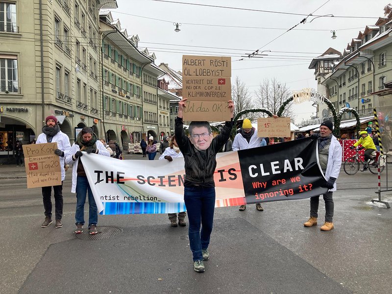 a protestor with a mask simulating albert rösti stands in front of a banner being held by slow marching scientists that reads the science is clear
